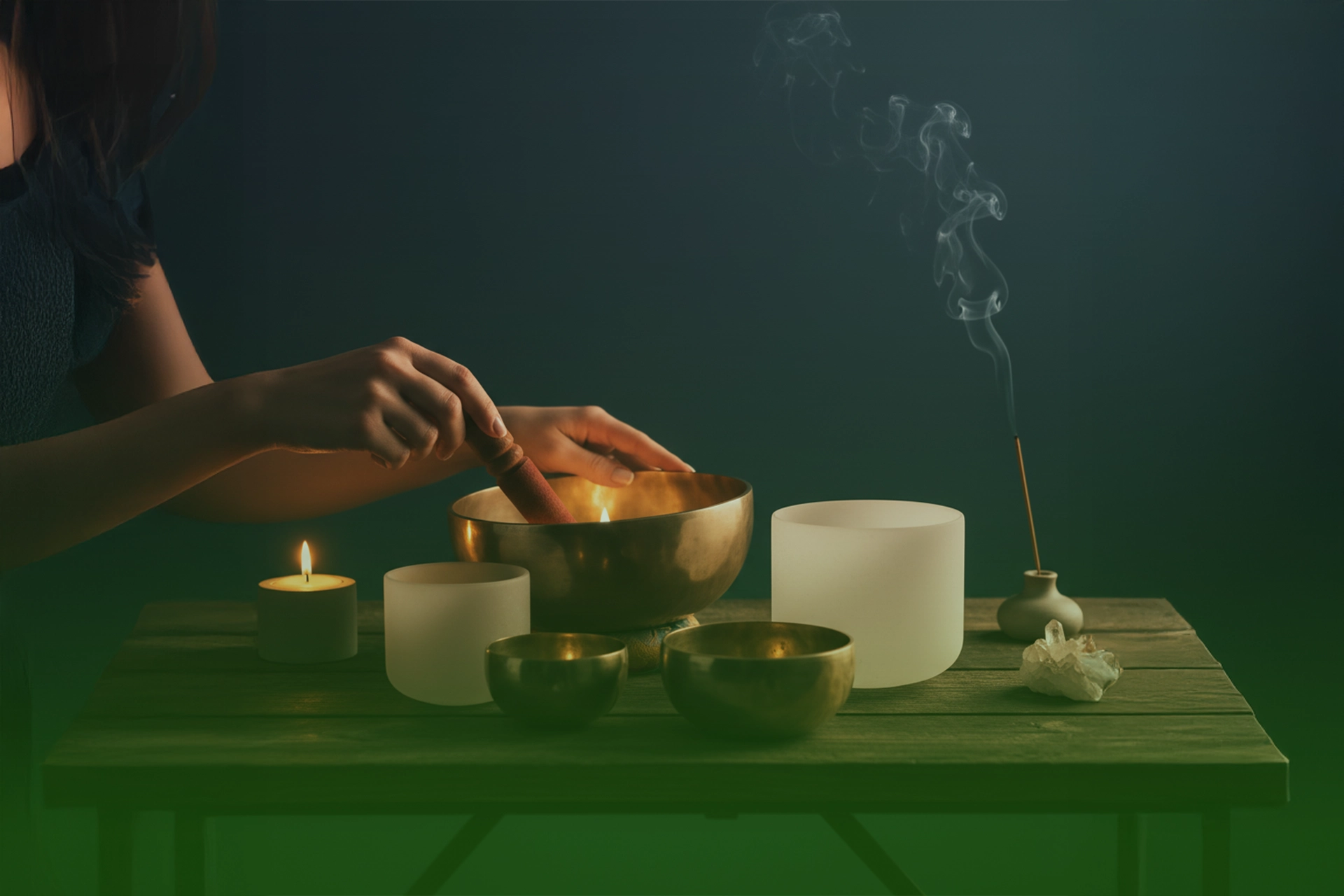 Manos haciendo sonar un cuenco tibetano de metal sobre una mesa con cuencos de cuarzo blancos, vela encendida e incienso humeante.