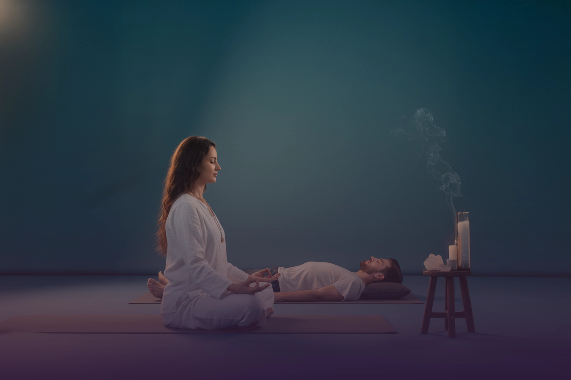 Guía de yoga en meditación junto a practicante en savasana, con velas, incienso humeante y cristales.