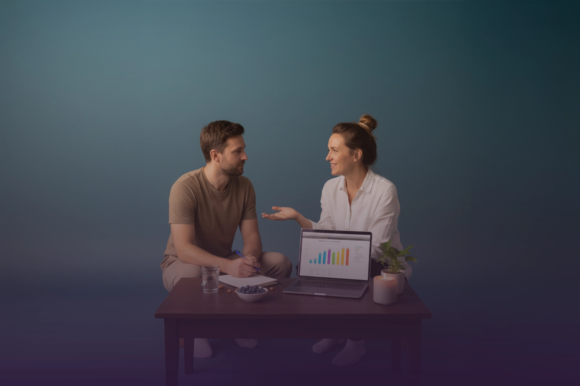 Coach conversa con cliente mientras revisan progreso en laptop; libreta, vaso de agua y vela sobre la mesa.